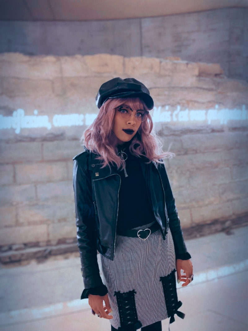 Aria Todd wearing a black cap, leather jacket, and plaid pencil skirt in front of neon lettering